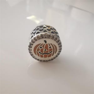 May include: A silver charm with a Halloween theme. The charm features a smiling jack-o'-lantern design in orange and black, surrounded by a white border and clear stones. The top of the charm has a pattern of small heart-shaped cutouts.