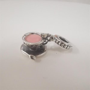 May include: A silver-coloured charm in the shape of a teacup and saucer, with a pink interior in the cup. The charm has an opening feature. The charm is detailed with decorative patterns and is designed to be a pendant.