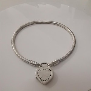 Può includere: Un braccialetto d'argento con un ciondolo a forma di lucchetto a forma di cuore. Il braccialetto ha un design a catena a serpente e un ciondolo a forma di cuore con una chiusura a lucchetto. Il ciondolo ha la scritta "5925 ALE".