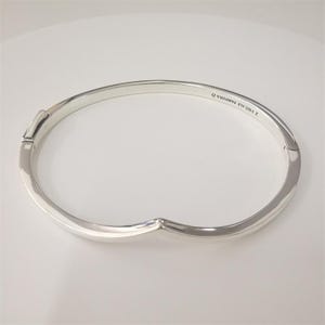 May include: A silver hinged bangle bracelet. The bracelet has a smooth, polished finish and a simple, elegant design. The clasp is visible on the left side. The bracelet is marked with "S 925 ALE PANDORA O".