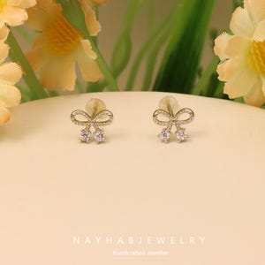 May include: Two silver stud earrings, each shaped like a bow with a clear gemstone at the base. The earrings are presented on a pale surface. The text "NAYHAB JEWELRY" and "Handcrafted Jeweller" are visible at the bottom of the image.