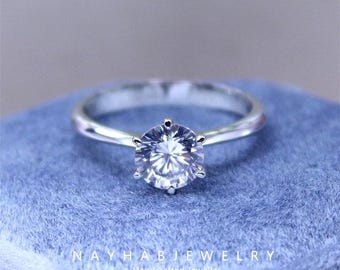 May include: A silver engagement ring with a large, clear, round-cut gemstone. The ring is set on a blue velvet surface. The text "NAYHAB JEWELRY Handcrafted Jeweller" is at the bottom of the image.