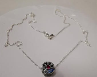 May include: A silver necklace with a pendant featuring a skull design. The skull pendant is adorned with red, blue, and black gemstones. The necklace is displayed on a white surface.