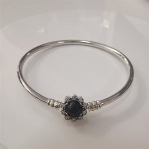 May include: A silver bangle bracelet featuring a black, flower-shaped charm. The charm is encircled by small, clear stones. The bracelet has a sleek, minimalist design.