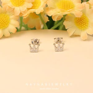 May include: A pair of silver crown stud earrings with clear gemstones. The earrings are displayed on a light beige surface, with yellow flowers in the background. The earrings are from NAYHABJEWELRY, Handcrafted Jeweller.