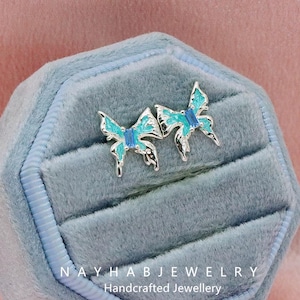 Puede incluir: Pendientes de mariposa plateados con esmalte turquesa y una gema azul rectangular, presentados en un joyero de terciopelo azul claro. Se aprecia el texto "NAYHAB JEWELRY Handcrafted Jewellery".