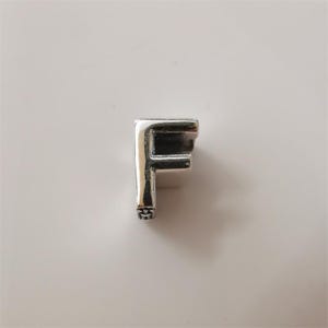 May include: A silver charm in the shape of the letter "F". The charm has a polished finish and a small decorative element at the bottom. The letter is displayed against a white background.