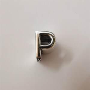 May include: A silver charm in the shape of the letter "P". The charm has a polished, reflective surface and a small decorative element at the bottom. The letter is in a sans-serif font.