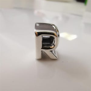 May include: A shiny silver charm in the shape of the letter "R". The charm has a polished, reflective surface and a hollowed-out center. The letter is in a serif font and stands upright.