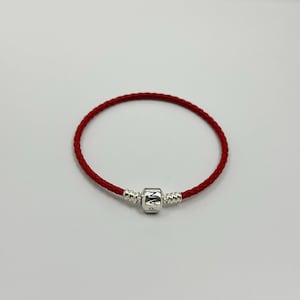 May include: A red braided bracelet with a silver clasp. The clasp is engraved with the letters "PAN". The bracelet is circular and rests on a white surface.