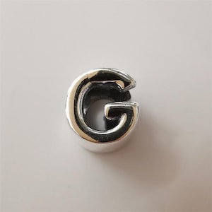 May include: A shiny, silver-colored charm in the shape of the letter "G". The charm has a rounded, three-dimensional design with a smooth, reflective surface. The letter is slightly open at the top.