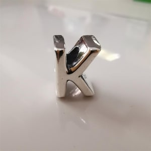 Puede incluir: Un charm de color plateado con forma de la letra "K". El charm tiene un acabado pulido y un pequeño lazo en la parte superior para sujetarlo a una pulsera o collar. La letra es de estilo bloque.