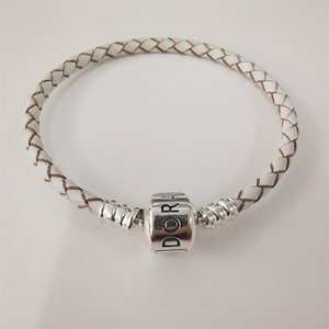 Op de afbeelding: Witte gevlochten leren armband met een zilveren bedel met de tekst "DORA". De armband heeft een geweven ontwerp met bruine accenten en zilveren accenten aan weerszijden van de bedel.