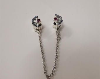 May include: Silver-toned charm with two skull-shaped components, each adorned with small blue and red gemstones. A delicate chain connects the two pieces. The charm is likely for a bracelet or necklace.
