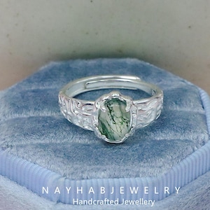 May include: A silver ring with an oval, green moss agate gemstone. The ring band has a textured, swirling pattern. The text "NAYHAB JEWELRY Handcrafted Jewellery" is at the bottom.