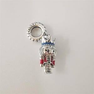 May include: A silver-colored charm with a nutcracker design. The charm features a red and silver uniform, a blue crown, and the word "PANDORA" on the loop. The charm is designed to be added to a bracelet or necklace.