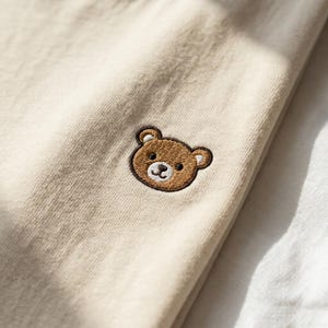 Puede incluir: Una prenda de tela color beige claro que presenta una peque&ntilde;a cara de oso bordada en color marr&oacute;n. El oso tiene orejas, ojos y nariz de color marr&oacute;n oscuro, con un hocico blanco y una sonrisa cosida sobre el tejido liso y uniforme.