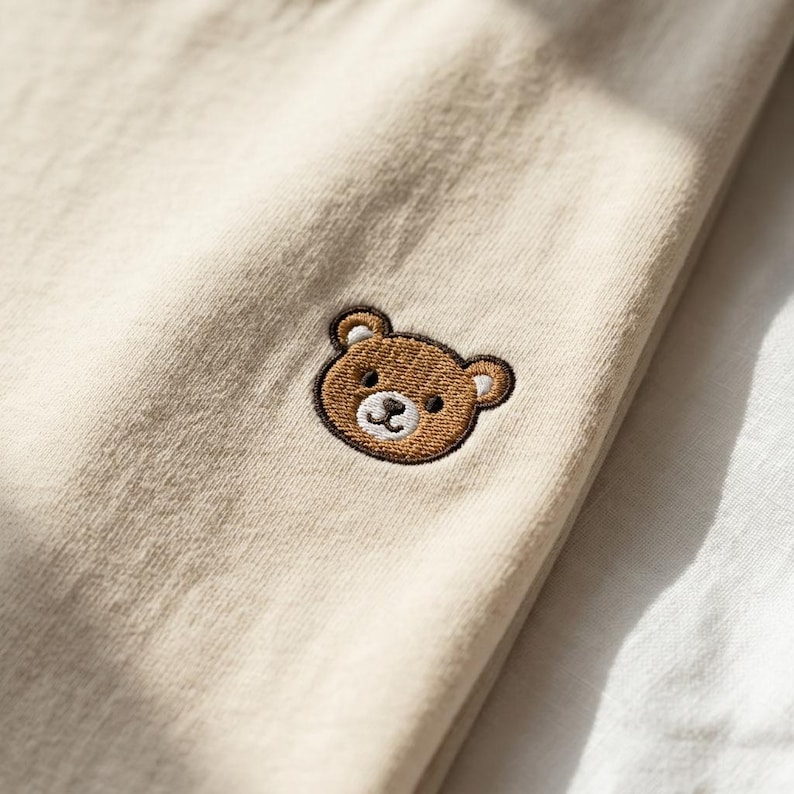 Puede incluir: Una prenda de tela color beige claro que presenta una peque&ntilde;a cara de oso bordada en color marr&oacute;n. El oso tiene orejas, ojos y nariz de color marr&oacute;n oscuro, con un hocico blanco y una sonrisa cosida sobre el tejido liso y uniforme.