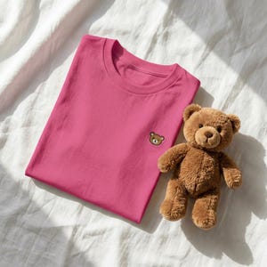 Puede incluir: Una camiseta de manga corta de color rosa brillante con una peque&ntilde;a cara de oso bordada en marr&oacute;n en el lado izquierdo del pecho, acompa&ntilde;ada de un oso de peluche marr&oacute;n con pelaje suave, orejas redondeadas y nariz cosida junto a la prenda.