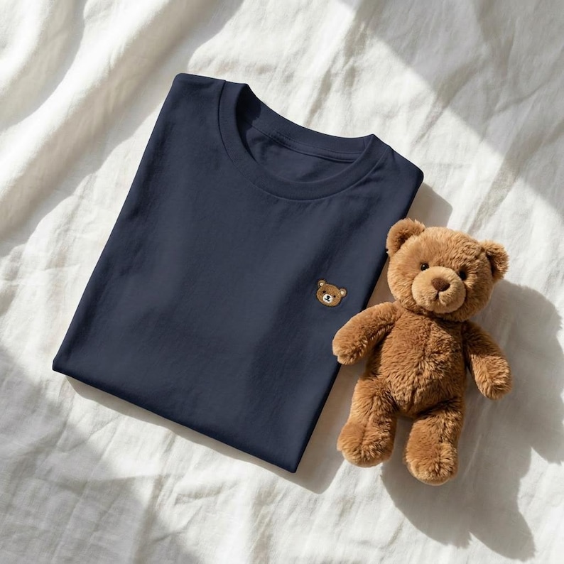 Puede incluir: Una camiseta azul marino con un peque&ntilde;o oso de peluche bordado en el lado izquierdo del pecho, colocada junto a un oso de peluche marr&oacute;n con ojos oscuros y nariz cosida, sobre una superficie de tela blanca.