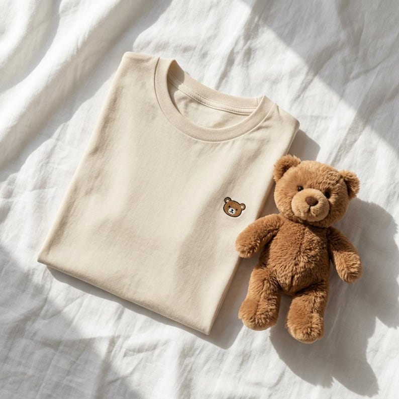 Puede incluir: Una camiseta de cuello redondo en color beige con una peque&ntilde;a cara de oso bordada en el lado izquierdo del pecho, acompa&ntilde;ada de un oso de peluche marr&oacute;n con pelaje suave, ojos oscuros y nariz cosida, situada junto a la prenda doblada.