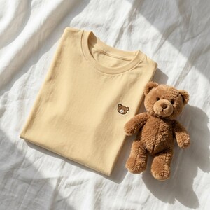 Puede incluir: Una camiseta de cuello redondo de color amarillo claro con una peque&ntilde;a cara de oso bordada en marr&oacute;n en el pecho izquierdo, acompa&ntilde;ada de un oso de peluche marr&oacute;n con ojos oscuros y nariz cosida, sobre una superficie de tela blanca.