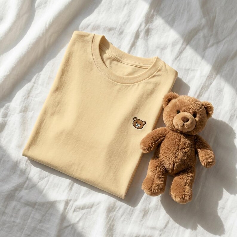 Puede incluir: Una camiseta de cuello redondo de color amarillo claro con una peque&ntilde;a cara de oso bordada en marr&oacute;n en el pecho izquierdo, acompa&ntilde;ada de un oso de peluche marr&oacute;n con ojos oscuros y nariz cosida, sobre una superficie de tela blanca.