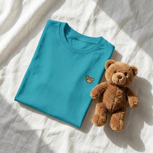Puede incluir: Una camiseta de cuello redondo color verde azulado con una peque&ntilde;a cara de oso marr&oacute;n bordada en el pecho izquierdo, colocada junto a un oso de peluche marr&oacute;n con pelaje suave, ojos oscuros y nariz cosida sobre una superficie de tela blanca.