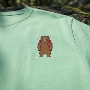 May include: A light green t-shirt featuring a cartoon bear with a red backpack. The bear is brown with a white and black face and is centered on the shirt. The shirt is made of a soft material.