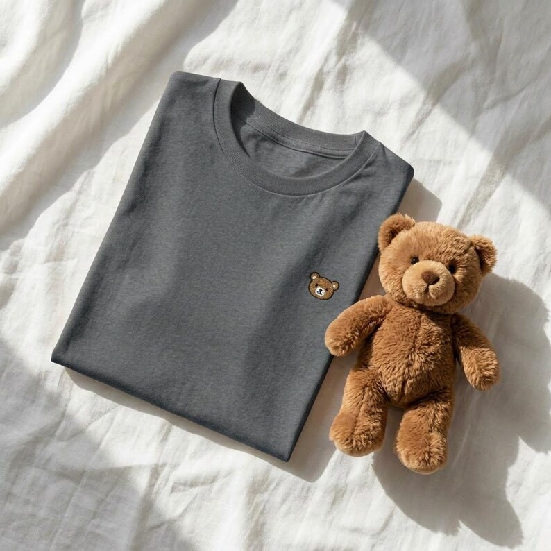 Puede incluir: Una camiseta gris carb&oacute;n de cuello redondo con una peque&ntilde;a cara de oso marr&oacute;n bordada en el pecho izquierdo, acompa&ntilde;ada de un oso de peluche marr&oacute;n con orejas redondeadas, nariz oscura y textura de pelaje suave junto a la prenda doblada.