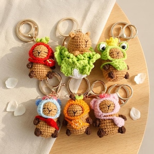 May include: A collection of six handmade crochet keychains. Each keychain features a small, brown animal figure with different colored hats and accessories, including a red hood, green lettuce, and a pink scarf. The keychains have gold-colored metal rings.