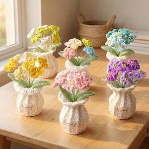 Pode incluir: Uma coleção de arranjos de flores de crochê feitos à mão em pequenos vasos brancos. As flores são de várias cores, incluindo amarelo, rosa, roxo e azul, com folhas verdes. Cada vaso tem uma pequena etiqueta que diz "handmade".