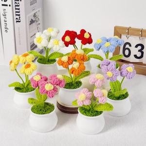 May include: A collection of handmade crochet flowers in various colors, including white, red, orange, yellow, pink, blue, and purple. Each flower is potted in a small white ceramic pot with green foliage.