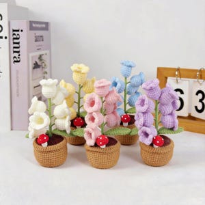 May include: A collection of six crochet flower arrangements in small woven baskets. Each arrangement features a different color of flowers, including white, yellow, pink, blue, and purple. Small red and white mushroom accents are included.