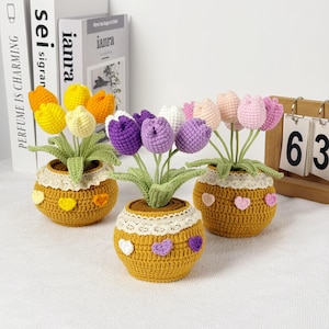 May include: Three crochet tulip flower arrangements in yellow pots. Each pot has a lace trim and heart accents. The tulips are in various colors, including yellow, orange, purple, white, and pink. The background includes books and a wooden calendar.