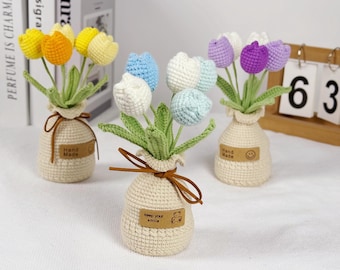 May include: Three crochet tulip bouquets in beige vases. The tulips are yellow, orange, white, blue, and purple. Each vase has a brown tag with text and a brown ribbon. The text on the tag says "Hand Made" and "keep your smile".