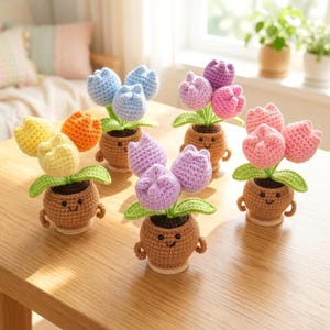 May include: A collection of handmade crochet tulip flowers in small brown pots with smiling faces. The tulips are in various colors, including yellow, orange, blue, purple, and pink. The pots have small arms and are sitting on a wooden table.