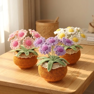 Puede incluir: Tres arreglos florales de ganchillo en macetas naranjas. Las flores son rosas, moradas y blancas con centros amarillos. Las macetas tienen un ribete de encaje. Los arreglos están sobre una mesa de madera.