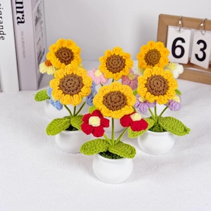 以下が含まれることがあります： 小さな白い鉢に入った手作りのかぎ針編みの花のコレクション。ヒマワリは黄色い花びらと茶色の中心部を持っています。他の花は赤、青、紫です。緑の葉と茎が花の配置を完成させます。木製のカレンダーには数字の63が表示されています。