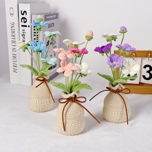 Peut inclure: Trois arrangements floraux au crochet dans des vases en tissu beige noués avec de la ficelle marron. Les fleurs sont dans des tons de bleu, rose, violet et blanc, avec des feuilles vertes. Le fond comprend des livres et un calendrier en bois.