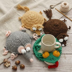 May include: Four crocheted animal-shaped coasters including a tan squirrel, a brown squirrel, a grey mole, and a green frog with a white ceramic mug placed on the frog coaster. Small decorative mushrooms and acorns are scattered on the surface.
