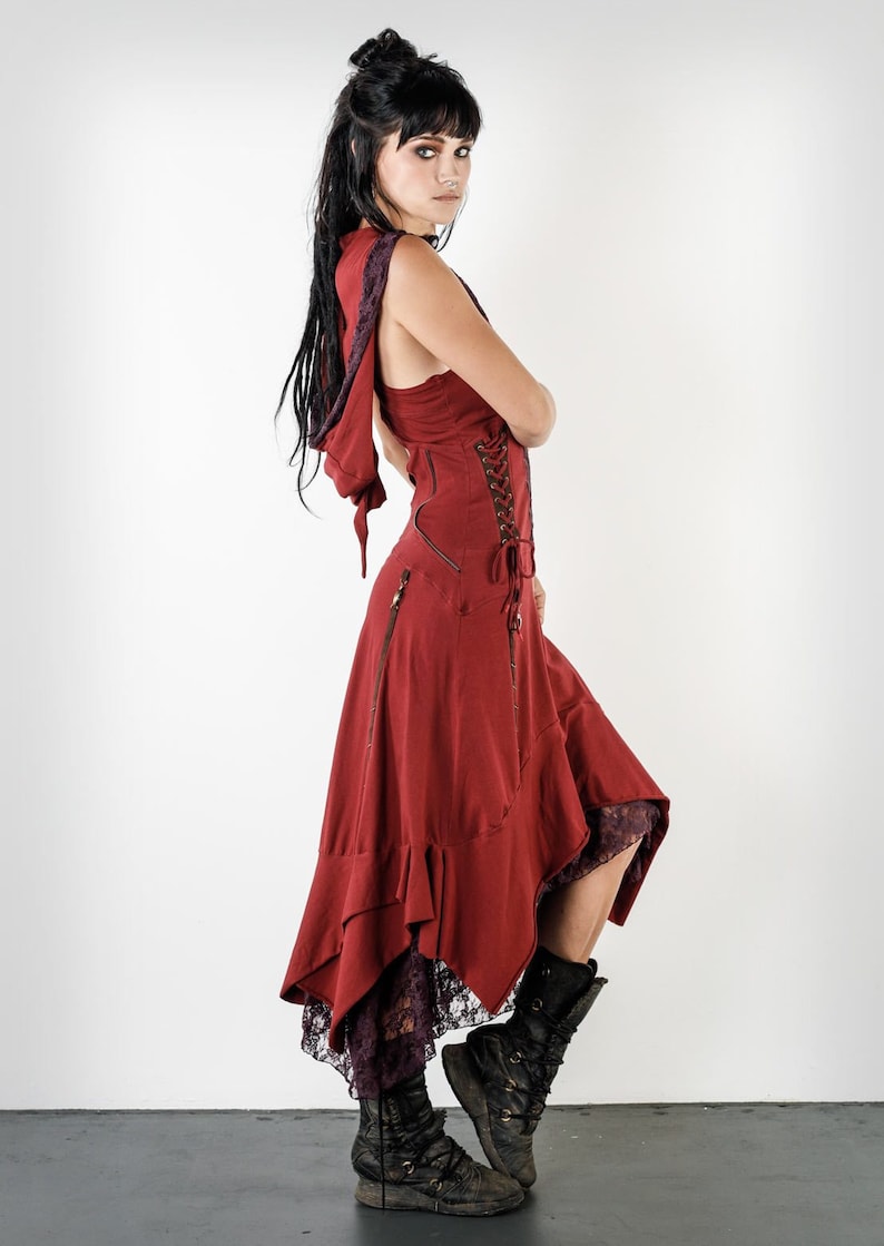 May include: A burgundy hooded dress with a lace trim and a lace-up back. The dress has a flowing skirt with a ruffled hem. The model is wearing black boots.