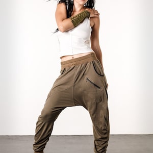 May include: A woman wearing a white tank top and brown, loose-fitting pants with a black waistband. She is wearing a green arm cuff on her left arm.