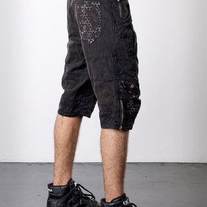 May include: Black cargo shorts with multiple pockets and zippers. The shorts feature a distressed look with a patterned design on the back pocket. The shorts are worn with black high-top sneakers.