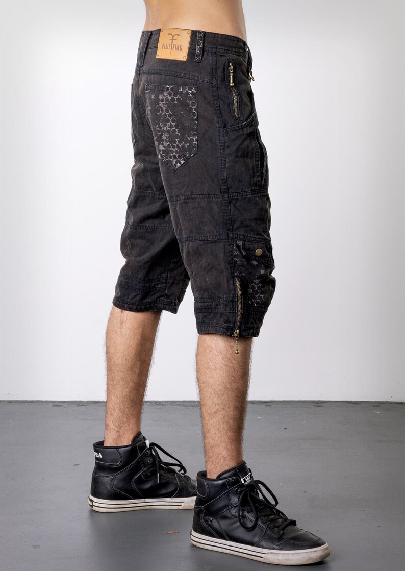 May include: Black cargo shorts with multiple pockets and zippers. The shorts feature a distressed look with a patterned design on the back pocket. The shorts are worn with black high-top sneakers.
