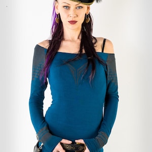 Könnte beinhalten: Eine Frau trägt ein blaues Langarmshirt mit einem schwarzen und goldenen Hut, einem schwarzen Gürtel und grauen Hosen mit einem schwarz-weißen Muster.