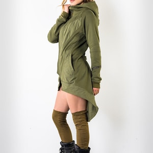 Könnte beinhalten: Eine langärmelige, olivgrüne Kapuzenjacke mit hohem Kragen und einem High-Low-Saum. Die Jacke hat einen Reißverschluss und zwei Taschen. Das Model trägt die Jacke mit schwarzen Stiefeln und braunen Kniestrümpfen.