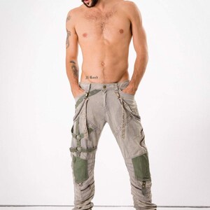 May include: A man wearing grey cargo pants with multiple straps and buckles. The pants have green fabric panels on the legs. The man is wearing white boots.