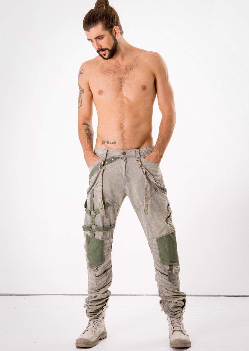 May include: A man wearing grey cargo pants with multiple straps and buckles. The pants have green fabric panels on the legs. The man is wearing white boots.