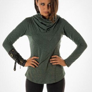 May include: A woman wearing a green long-sleeved shirt with a cowl neck and black pants. She is wearing a black leather cuff with metal accents on her left wrist.